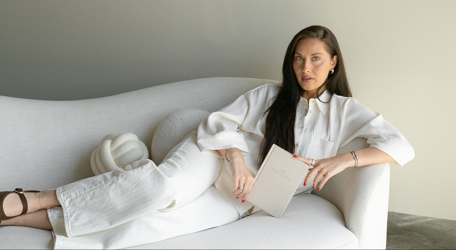 Woman sitting on a white couch holding a book in a minimalistic room.